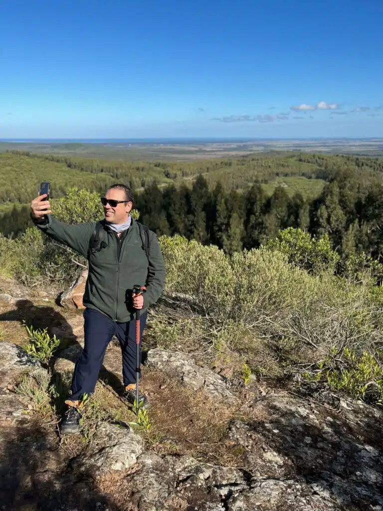 Cristhian de Mateando Rumbos tomando una selfie en un sendero, un claro ejemplo del disfrute del turismo sostenible con la naturaleza y el mar de fondo.